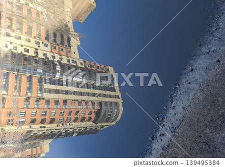 A modern brick residential building stands tall, its reflection visible in a puddle on the street. Rainwater creates a clear view of the structure as daylight shines. A modern brick residential building stands tall, its reflection visible in a puddle on the street. Rainwater creates a clear view of the structure as daylight shines. 139495384