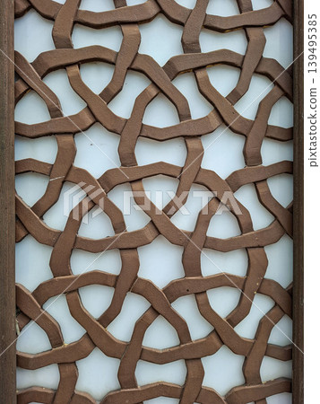 A detailed view shows a brown wooden lattice with an intricate design of interlocking shapes. The pattern is based on Islamic art, set against a bright white background. A detailed view shows a brown wooden lattice with an intricate design of interlocking shapes. The pattern is based on Islamic art, set against a bright white background. 139495385