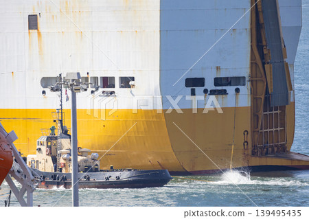 Large cargo ship is being loaded while a tugboat assists near the port during bright daylight hours in a busy harbor Large cargo ship is being loaded while a tugboat assists near the port during bright daylight hours in a busy harbor 139495435