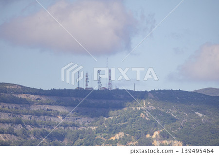 Communication towers stand on a mountain during the day with clouds in the sky and greenery on the slopes visible 139495464