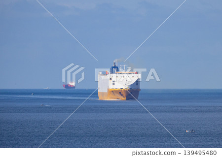 Cargo ship sails over calm waters near coastline in bright daylight with smaller boats in the background Cargo ship sails over calm waters near coastline in bright daylight with smaller boats in the background 139495480
