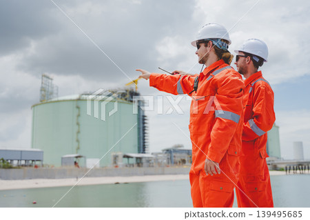 Engineers in orange uniforms inspecting LNG storage tanks at coastal terminal under cloudy sky 139495685