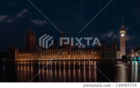 Illuminated clock tower and long gothic building stand beside river under dark sky. Warm lights reflect on water forming clear architectural night scene. Illuminated clock tower and long gothic building stand beside river under dark sky. Warm lights reflect on water forming clear architectural night scene. 139495714