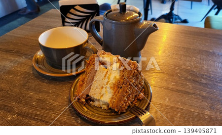A slice of fresh cake on a plate next to a cup of hot tea on a wooden table, cozy cafe morning A slice of fresh cake on a plate next to a cup of hot tea on a wooden table, cozy cafe morning 139495871