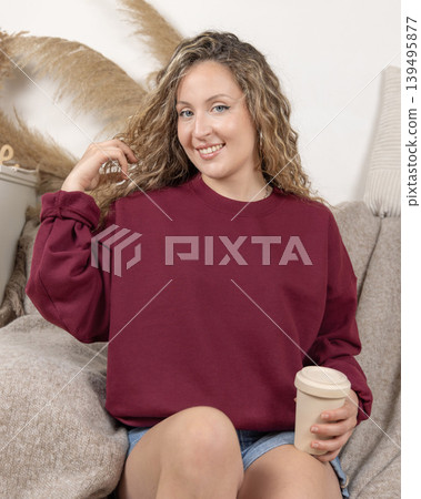 Blank maroon oversized sweatshirt mockup on fair-haired young woman sitting on sofa, front view Blank maroon oversized sweatshirt mockup on fair-haired young woman sitting on sofa, front view 139495877