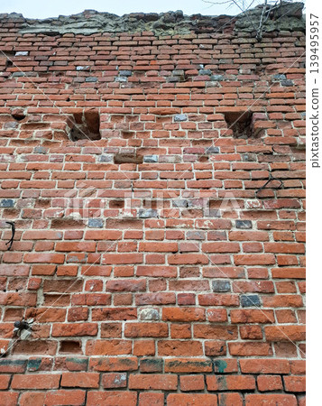 A red brick wall stands worn and damaged. The bricks show a rough texture, and the mortar is crumbling. There are holes and voids scattered across the surface. A red brick wall stands worn and damaged. The bricks show a rough texture, and the mortar is crumbling. There are holes and voids scattered across the surface. 139495957