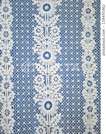Intricate white lace with floral designs and symmetrical embroidery set against a bright blue background. The lace is handmade and showcases detailed craftsmanship. Intricate white lace with floral designs and symmetrical embroidery set against a bright blue background. The lace is handmade and showcases detailed craftsmanship. 139495962