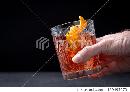 Hand holding classic old fashioned cocktail with orange garnish on dark background Hand holding classic old fashioned cocktail with orange garnish on dark background 139495973