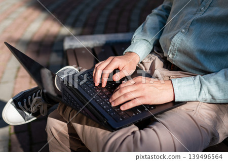 Man sitting on bench and working on laptop. Freelancer typing on keyboard in park. Digital nomad lifestyle, remote work and business communication in urban city environment concept. 139496564