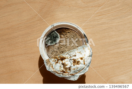 Moldy food in small plastic container with opened foil lid. Spoiled product on table surface. Unsafe nutrition and hygiene risk concept. Expired meal waste and health hazard. Moldy food in small plastic container with opened foil lid. Spoiled product on table surface. Unsafe nutrition and hygiene risk concept. Expired meal waste and health hazard. 139496567