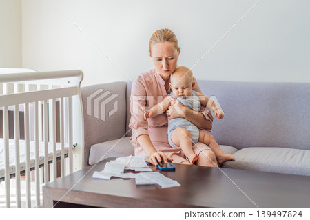 Mother sitting on sofa holding baby while calculating monthly expenses with receipts at home. Stressed mom managing budget and childcare. Financial stress, parenthood and real life concept Mother sitting on sofa holding baby while calculating monthly expenses with receipts at home. Stressed mom managing budget and childcare. Financial stress, parenthood and real life concept 139497824
