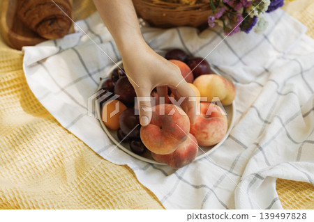 Hand reaching for a fresh peach from a fruit plate on a summer picnic blanket outdoors Hand reaching for a fresh peach from a fruit plate on a summer picnic blanket outdoors 139497828