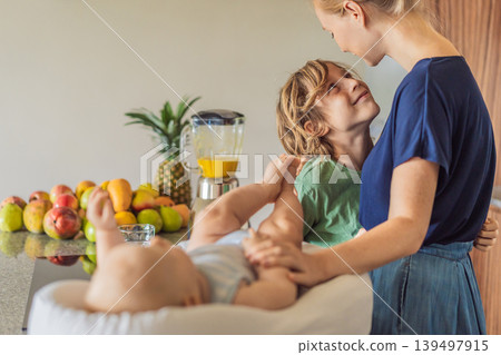 Mother and son preparing smoothie in kitchen while baby lies in cozy lounger on table watching. Family cooking together, healthy lifestyle, parenting, sibling bonding and everyday life concept Mother and son preparing smoothie in kitchen while baby lies in cozy lounger on table watching. Family cooking together, healthy lifestyle, parenting, sibling bonding and everyday life concept 139497915
