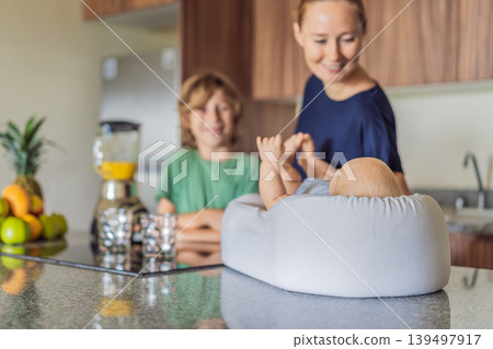 Mother and son preparing smoothie in kitchen while baby lies in cozy lounger on table watching. Family cooking together, healthy lifestyle, parenting, sibling bonding and everyday life concept Mother and son preparing smoothie in kitchen while baby lies in cozy lounger on table watching. Family cooking together, healthy lifestyle, parenting, sibling bonding and everyday life concept 139497917