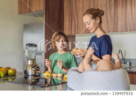 Mother and son preparing smoothie in kitchen while baby lies in cozy lounger on table watching. Family cooking together, healthy lifestyle, parenting, sibling bonding and everyday life concept Mother and son preparing smoothie in kitchen while baby lies in cozy lounger on table watching. Family cooking together, healthy lifestyle, parenting, sibling bonding and everyday life concept 139497918