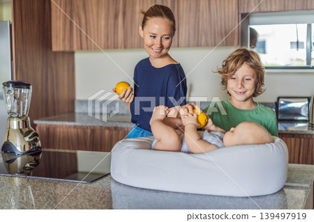 Mother and son preparing smoothie in kitchen while baby lies in cozy lounger on table watching. Family cooking together, healthy lifestyle, parenting, sibling bonding and everyday life concept Mother and son preparing smoothie in kitchen while baby lies in cozy lounger on table watching. Family cooking together, healthy lifestyle, parenting, sibling bonding and everyday life concept 139497919
