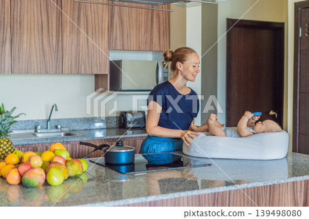 Mother talking to baby lying in cozy lounger on kitchen table while cooking at home. Caring mom interacting with infant during daily routine. Parenting, multitasking and family life concept Mother talking to baby lying in cozy lounger on kitchen table while cooking at home. Caring mom interacting with infant during daily routine. Parenting, multitasking and family life concept 139498080