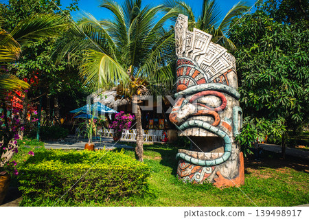 A large tiki totem statue stands in a tropical garden with palm trees. A large tiki totem statue stands in a tropical garden with palm trees. 139498917
