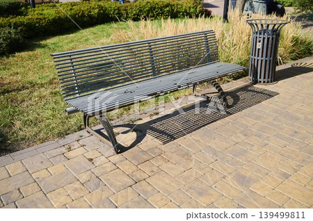 Sunlit park scene showcasing a metal bench casting shadows on stone pavement Sunlit park scene showcasing a metal bench casting shadows on stone pavement 139499811