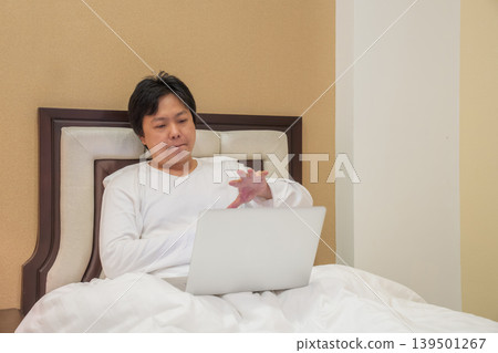 Man sits on bed using laptop while raising hand toward screen, combining remote work lifestyle with touchless gesture control and digital interaction. Man sits on bed using laptop while raising hand toward screen, combining remote work lifestyle with touchless gesture control and digital interaction. 139501267