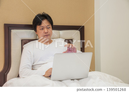 Man sits on bed using laptop while raising hand toward screen, combining remote work lifestyle with touchless gesture control and digital interaction. Man sits on bed using laptop while raising hand toward screen, combining remote work lifestyle with touchless gesture control and digital interaction. 139501268
