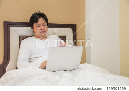 Man sits on bed using laptop while raising hand toward screen, combining remote work lifestyle with touchless gesture control and digital interaction. Man sits on bed using laptop while raising hand toward screen, combining remote work lifestyle with touchless gesture control and digital interaction. 139501269