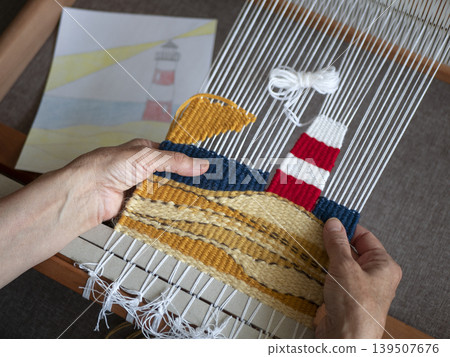 Close-up of a person weaving a handmade tapestry with lighthouse motif on a wooden frame loom. Weaver following a paper sketch to create a textile landscape with colorful wool yarns Close-up of a person weaving a handmade tapestry with lighthouse motif on a wooden frame loom. Weaver following a paper sketch to create a textile landscape with colorful wool yarns 139507676