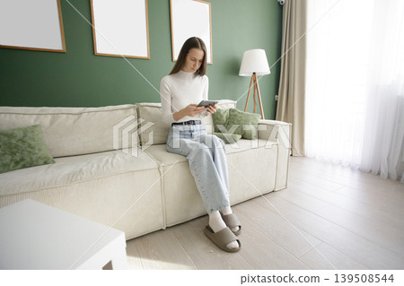 Lifestyle portrait of woman reading ebook gadget sitting on comfortable sofa in modern interior Lifestyle portrait of woman reading ebook gadget sitting on comfortable sofa in modern interior 139508544