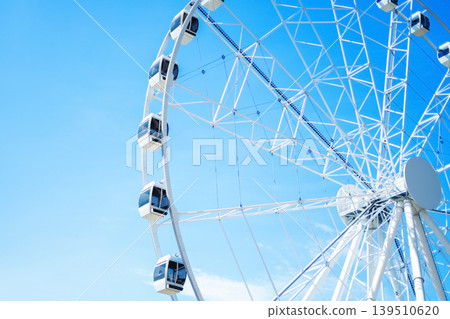 A large white Ferris wheel with modern enclosed gondolas set against a vibrant, clear blue sky. A minimalist and bright scene capturing summer fun and amusement park vibes. 139510620