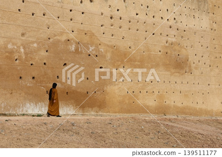 Women walks beside a massive, textured earthen wall 139511177