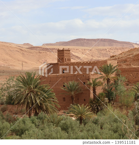 A traditional earthen kasbah with a square tower built from mud bricks rises from a lush oasis surrounded by palm trees in a vast arid desert landscape. A traditional earthen kasbah with a square tower built from mud bricks rises from a lush oasis surrounded by palm trees in a vast arid desert landscape. 139511181