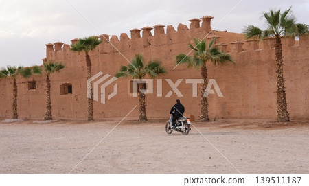 A lone motorcyclist rides past a traditional adobe fortress lined with palm trees in a desert setting A lone motorcyclist rides past a traditional adobe fortress lined with palm trees in a desert setting 139511187