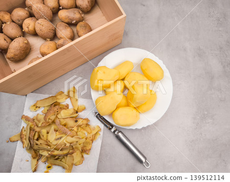 Peeled potatoes on plate, peels, peeler and wooden crate on kitchen counter, copy space 139512114