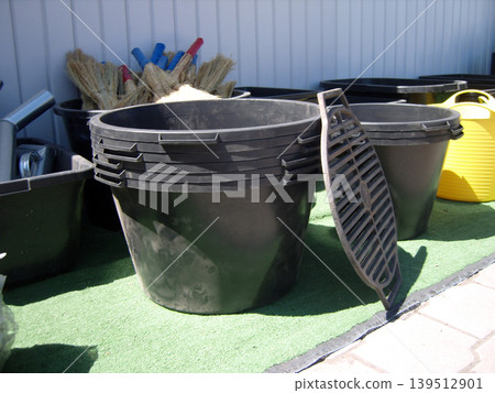 Neat outdoor arrangement of black plastic tubs, colorful buckets, and traditional straw brooms on green turf. Essential utility gear for gardening, household chores, and organized yard storage Neat outdoor arrangement of black plastic tubs, colorful buckets, and traditional straw brooms on green turf. Essential utility gear for gardening, household chores, and organized yard storage 139512901