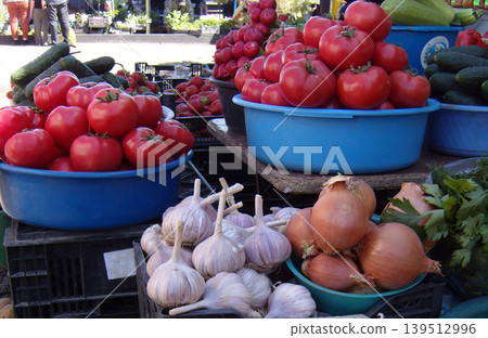 Vibrant outdoor market stall overflowing with fresh summer produce. Plump red tomatoes in blue basins take center stage, accompanied by crisp cucumbers, pungent garlic bulbs, and golden onions Vibrant outdoor market stall overflowing with fresh summer produce. Plump red tomatoes in blue basins take center stage, accompanied by crisp cucumbers, pungent garlic bulbs, and golden onions 139512996