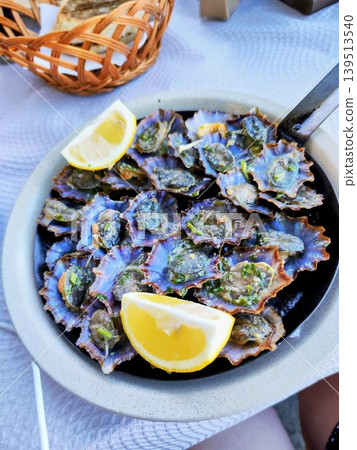 A dish featuring several grilled limpets, Patella spp. served in a round ceramic bowl, garnished with two lemon wedges. A woven basket with slices of bread A dish featuring several grilled limpets, Patella spp. served in a round ceramic bowl, garnished with two lemon wedges. A woven basket with slices of bread 139513540