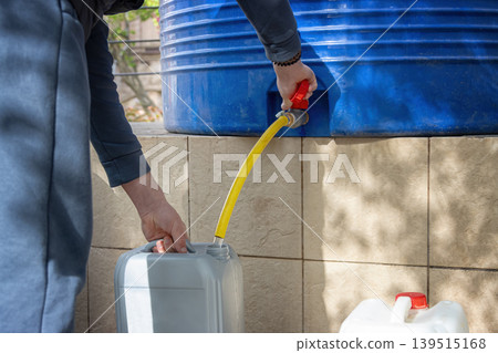 Person filling plastic jerrycan with water from large blue storage tank outdoors 139515168