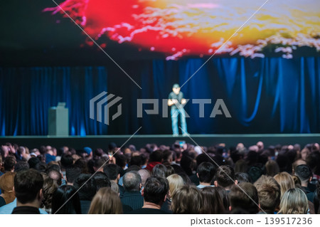 Conference speaker on stage presenting to large audience under colorful screen during tech event Conference speaker on stage presenting to large audience under colorful screen during tech event 139517236