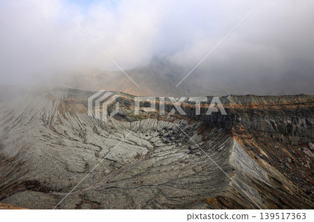 View of the crater opening of erupting volcano. Crater with volcanic gas fumaroles. Clouds and steam in the sky.  139517363