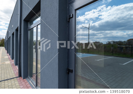 A modern building shows clear glass windows reflecting clouds, with an empty parking lot beside it under a blue sky 139518070