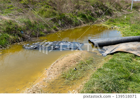A stream shows brown water and trash along the bank. Plastic and rubber materials collect by the water's edge in spring sunlight A stream shows brown water and trash along the bank. Plastic and rubber materials collect by the water's edge in spring sunlight 139518071