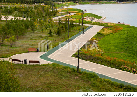 Embankment of a river with a walking path through a park. Scenic landscape design with benches and trees by the water. Recreational public space for city leisure and relaxation. Embankment of a river with a walking path through a park. Scenic landscape design with benches and trees by the water. Recreational public space for city leisure and relaxation. 139518120