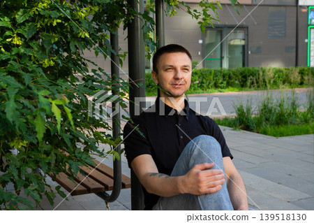 Man resting on wooden bench in city park. Caucasian guy sitting outdoors near green tree. Leisure time in urban environment, peaceful break from daily life, summer lifestyle concept. Man resting on wooden bench in city park. Caucasian guy sitting outdoors near green tree. Leisure time in urban environment, peaceful break from daily life, summer lifestyle concept. 139518130