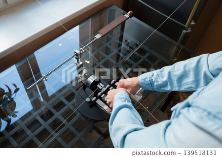 Man adjusting professional knife sharpener on glass table. Master setting up manual metal sharpening tool for kitchen blade maintenance. Craftsmanship and home utility equipment concept. Man adjusting professional knife sharpener on glass table. Master setting up manual metal sharpening tool for kitchen blade maintenance. Craftsmanship and home utility equipment concept. 139518131