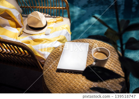 Cozy outdoor setting featuring a wicker chair with a yellow striped blanket, a sun hat, a blank notebook, and a cup of coffee beside a swimming pool 139518747