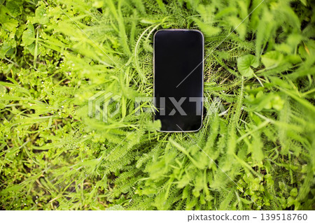 Smartphone lying on lush green grass surrounded by vibrant foliage, showcasing a modern device in a natural outdoor setting Smartphone lying on lush green grass surrounded by vibrant foliage, showcasing a modern device in a natural outdoor setting 139518760