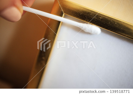 Close-up of a hand using a cotton swab to clean the edge of a gold-framed picture, highlighting the cleaning process and attention to detail in home decor maintenance Close-up of a hand using a cotton swab to clean the edge of a gold-framed picture, highlighting the cleaning process and attention to detail in home decor maintenance 139518771