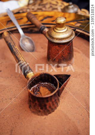 Traditional coffee brewing equipment made of metal sits on fine sand, showcasing intricate designs and textures, highlighting the art of coffee preparation and cultural significance Traditional coffee brewing equipment made of metal sits on fine sand, showcasing intricate designs and textures, highlighting the art of coffee preparation and cultural significance 139518814