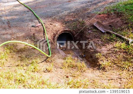 Underground pipe opening near dirt path with green hose nearby tools scattered Underground pipe opening near dirt path with green hose nearby tools scattered 139519015