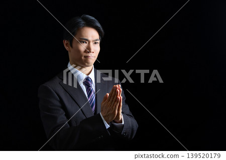 A businessman in a suit praying with his hands clasped together against a black background. 139520179
